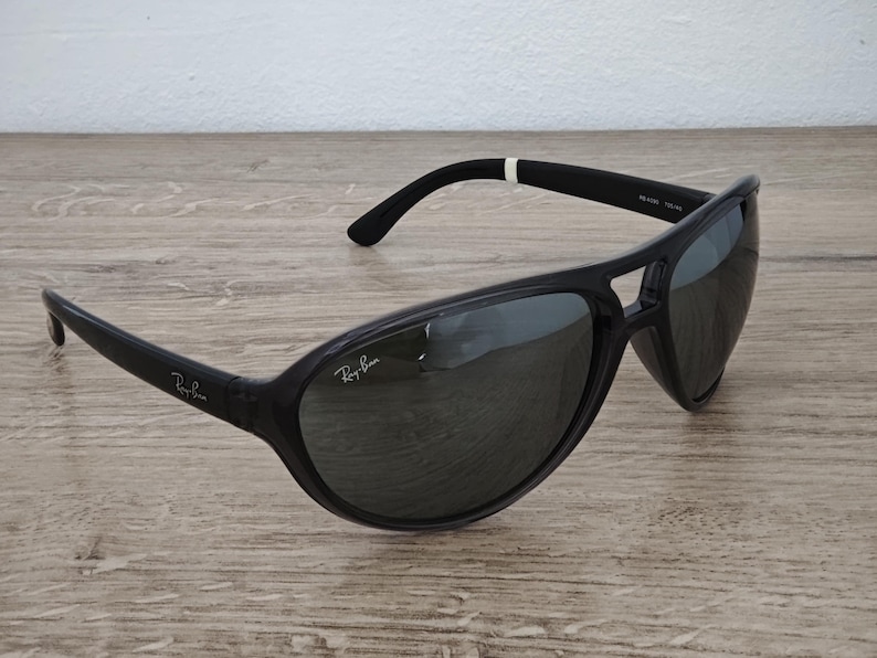 Puede incluir: Gafas de sol negras con lentes oscuras. El logotipo de Ray-Ban es visible en las patillas. Las gafas de sol tienen un dise&ntilde;o deportivo y envolvente. La montura es negra y las lentes son de color gris oscuro. Est&aacute;n sobre una superficie de madera.