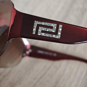 May include: Close-up of burgundy sunglasses with a silver geometric design on the temple. The lenses are a gradient brown color. The sunglasses are on a light brown surface.