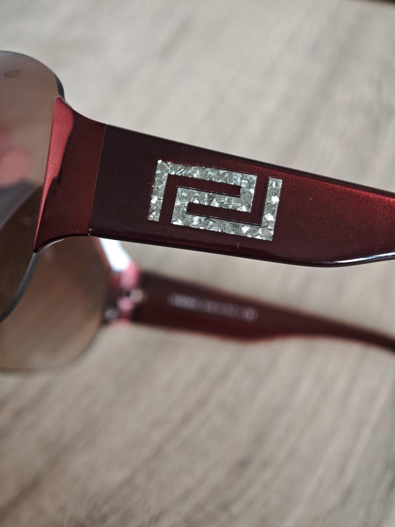 May include: Close-up of burgundy sunglasses with a silver geometric design on the temple. The lenses are a gradient brown color. The sunglasses are on a light brown surface.