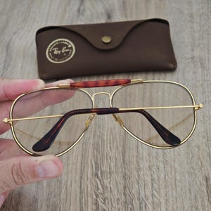 K&ouml;nnte beinhalten: Goldgerahmte Aviator-Brille mit klaren Gl&auml;sern und braunen B&uuml;geln. Die Brille wird mit einem braunen Lederetui geliefert. Das Ray-Ban-Logo ist sichtbar.