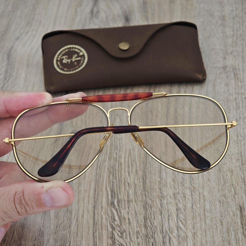 K&ouml;nnte beinhalten: Goldgerahmte Aviator-Brille mit klaren Gl&auml;sern und braunen B&uuml;geln. Die Brille wird mit einem braunen Lederetui geliefert. Das Ray-Ban-Logo ist sichtbar.