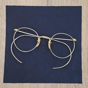 May include: Gold-toned metal eyeglasses featuring round frames and long, curved cable temples. The eyewear includes a thin bridge, adjustable nose pads, and circular rims designed to hold lenses, showcasing a classic wire-frame construction.