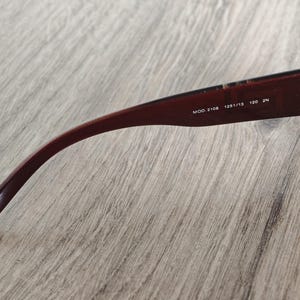 May include: Close-up of the arm of a pair of sunglasses. The arm is a deep burgundy color with the text "MOD. 2108 125/13 120 2N" printed in white. The sunglasses are resting on a light brown wooden surface.