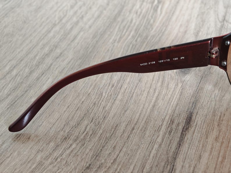 May include: Close-up of the arm of a pair of sunglasses. The arm is a deep burgundy color with the text "MOD. 2108 125/13 120 2N" printed in white. The sunglasses are resting on a light brown wooden surface.