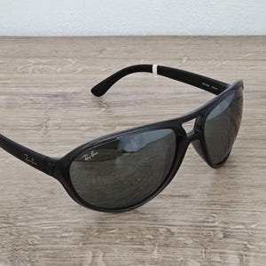 Puede incluir: Gafas de sol Ray-Ban negras con un dise&ntilde;o deportivo. Las gafas tienen una montura curvada y lentes oscuras. El logotipo de Ray-Ban es visible en la lente y la patilla izquierda. Sobre una superficie de madera clara.