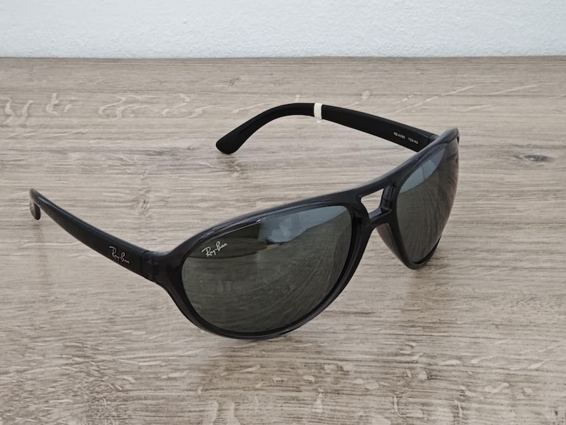 Puede incluir: Gafas de sol Ray-Ban negras con un dise&ntilde;o deportivo. Las gafas tienen una montura curvada y lentes oscuras. El logotipo de Ray-Ban es visible en la lente y la patilla izquierda. Sobre una superficie de madera clara.