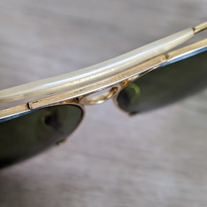 K&ouml;nnte beinhalten: Nahaufnahme einer goldgerahmten Sonnenbrille mit gr&uuml;nen Gl&auml;sern. Der Rahmen hat ein geschwungenes Design mit einem hellen B&uuml;gel. Der Text "RAY-BAN 1/10 12K GF" ist auf dem Rahmen zu sehen.