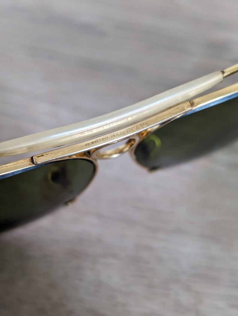 K&ouml;nnte beinhalten: Nahaufnahme einer goldgerahmten Sonnenbrille mit gr&uuml;nen Gl&auml;sern. Der Rahmen hat ein geschwungenes Design mit einem hellen B&uuml;gel. Der Text "RAY-BAN 1/10 12K GF" ist auf dem Rahmen zu sehen.