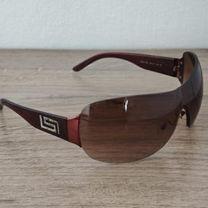 May include: Brown sunglasses with a wraparound design. The lenses are a gradient brown, and the frames are a dark brown with a decorative silver emblem on the temples. The sunglasses are set against a wooden surface.