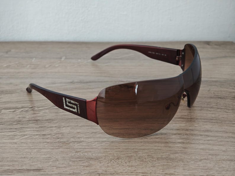 May include: Brown sunglasses with a wraparound design. The lenses are a gradient brown, and the frames are a dark brown with a decorative silver emblem on the temples. The sunglasses are set against a wooden surface.