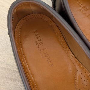 May include: Close-up of a pair of brown leather loafers. The interior of one shoe is visible, displaying the embossed text "RALPH LAUREN MADE IN U.S.A." on a tan insole. The shoe's edges are a darker brown.