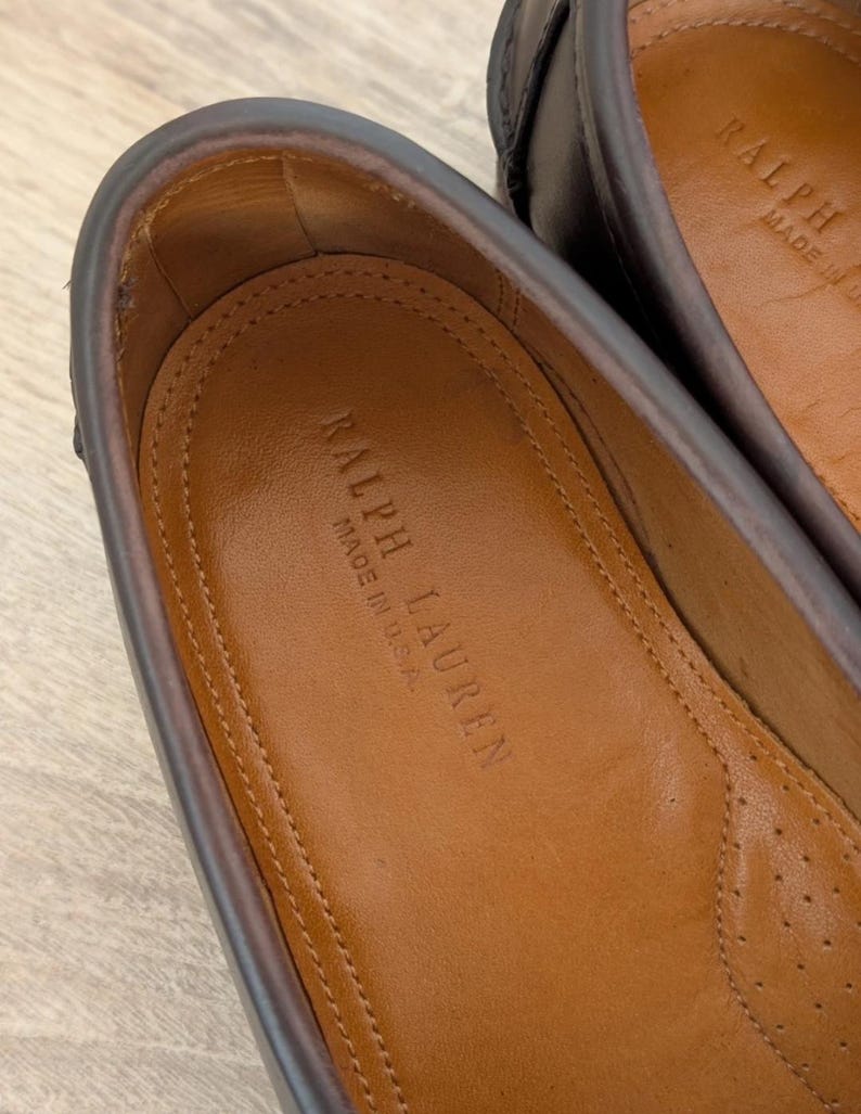 May include: Close-up of a pair of brown leather loafers. The interior of one shoe is visible, displaying the embossed text "RALPH LAUREN MADE IN U.S.A." on a tan insole. The shoe's edges are a darker brown.
