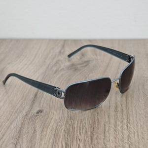 Puede incluir: Gafas de sol azuladas con lentes marrones degradadas. Las gafas tienen una montura rectangular y el logo de Chanel en las patillas. Las varillas son de color azul oscuro.