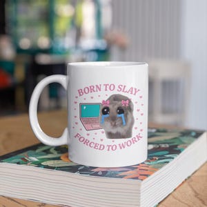 May include: A white ceramic mug featuring a graphic of a crying hamster with pink bows, a pink laptop, and the text BORN TO SLAY FORCED TO WORK surrounded by small pink hearts. The design is printed on the front of the mug with a handle on the left.