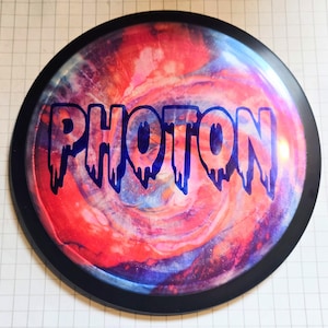 Könnte beinhalten: Eine kreisförmige Disc-Golf-Scheibe mit einem lebendigen, wirbelnden Batikmuster in Rot-, Rosa- und Blautönen. Das Wort PHOTON ist in der Mitte in einer fetten, dunkelblauen Schrift mit einem markanten Tropf- oder Schmelzeffekt aufgedruckt.