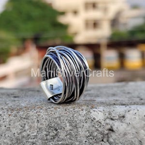 Puede incluir: Un anillo de plata con un diseño de alambre tejido. El anillo presenta múltiples hebras de alambre de plata enrolladas entre sí, creando un efecto texturizado y en capas. El anillo tiene un acabado pulido y una pequeña banda rectangular con el texto "925".