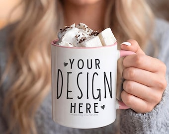 May include: A white mug with a pink handle and rim, filled with whipped cream, marshmallows, and chocolate shavings. The mug has the text "YOUR DESIGN HERE" in black. The mug is held in a person's hand.