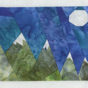 May include: A fabric art piece depicting a mountain landscape with a night sky. The mountains are green and blue, with white peaks. The sky is a gradient of blues, and a white, geometric moon is in the upper right corner.