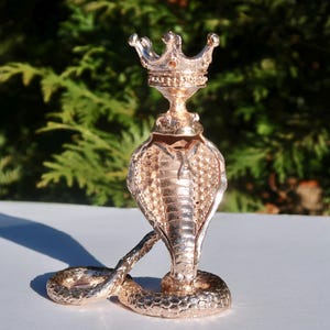 May include: A detailed, metallic cobra figurine with a crown atop its head. The cobra is in a striking pose, with its hood flared and its body coiled. The figurine is a bronze color and sits on a white surface, with a green background.