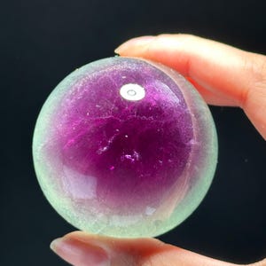 Puede incluir: Una esfera translúcida, principalmente morada en el centro, que transiciona a un tono verde pálido hacia los bordes. La bola de cristal pulida se sostiene entre los dedos sobre un fondo oscuro. La esfera tiene una superficie lisa y reflectante.