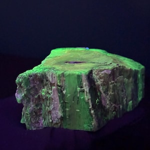 May include: A fluorescent green mineral specimen with a rough, textured surface. The rock has a rectangular shape with a jagged edge, and the color is a vibrant, glowing green. The background is dark.