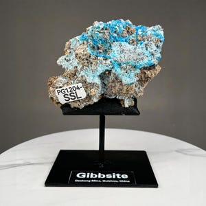 May include: A specimen of Gibbsite, a mineral, displayed on a black stand. The mineral features a mix of light blue and brown colors. A small white label with the text "PG1204-SSL" is attached to the mineral. The base of the stand has the text "Gibbsite" and "Dachang Mine, Guizhou, China".