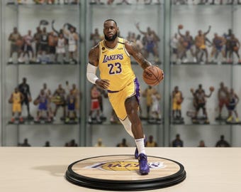 May include: A figurine of a basketball player in a yellow and purple jersey with the number 23, dribbling a basketball. The figurine is on a wooden base with the Lakers logo and a black border. Other figurines are in the background.