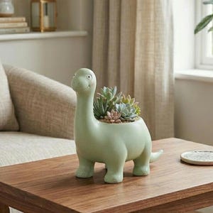 May include: A light green dinosaur-shaped planter filled with succulents sits on a wooden table. The planter has a long neck, a smiling face, and short legs. The succulents are a mix of green, pink, and gray.