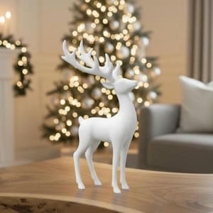 Könnte beinhalten: Eine weiße dekorative Rentierfigur mit großem Geweih steht auf einem Holztisch. Das Rentier steht vor einem Weihnachtsbaum mit leuchtenden Lichtern und schafft eine festliche Urlaubsszene.
