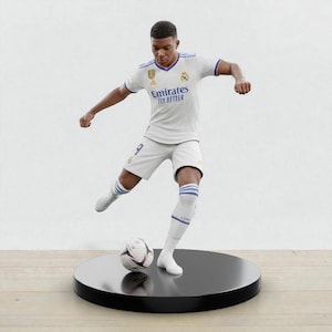 Könnte beinhalten: Figur einer Fußballspielers in weißer Uniform mit blauen Akzenten, der einen Fußball tritt. Die Figur steht auf einem schwarzen, kreisförmigen Sockel. Auf der Uniform steht "Emirates Fly Better".