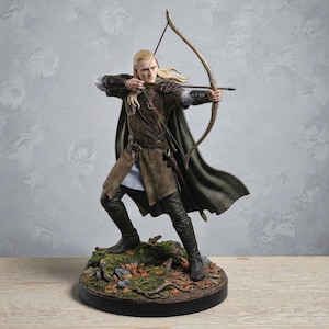 May include: A detailed figurine of an archer in a brown tunic, cape, and black boots, drawing a bow. The figure has long blonde hair and is posed on a base with foliage and a black border. The overall theme is fantasy.