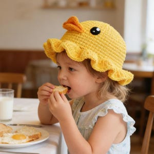 Può includere: Un bambino indossa un cappello da anatra giallo all'uncinetto con becco arancione e occhio nero. Il cappello ha un bordo a balze. Il bambino sta mangiando pane a tavola con un piatto di cibo, un bicchiere di latte e posate.