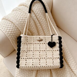 Peut inclure: Sac fourre-tout en crochet crème avec garniture et poignées noires. Le sac présente un motif ajouré et un petit charme en forme de cœur noir. Une petite étiquette métallique est attachée à l'avant du sac.
