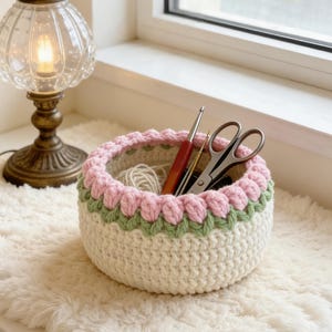 May include: A handmade crochet basket in cream, with a pink and green scalloped border. The basket contains yarn, a crochet hook, and scissors. The basket sits on a fluffy white surface near a window and a vintage-style lamp.