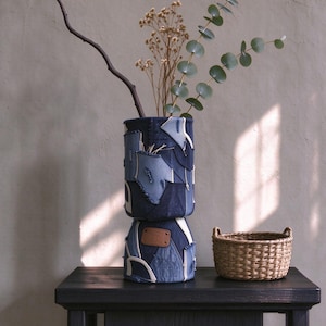 May include: A cylindrical vase constructed from various shades of blue denim fabric patches with visible stitching and a brown leather label. The vase contains a bare branch, dried flowers, and a green eucalyptus stem.