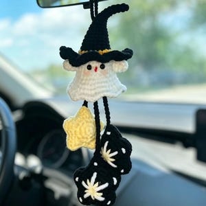 May include: A handmade crochet car charm featuring a white ghost wearing a black witch hat with a yellow brim. Below the ghost are yellow stars and black star-shaped accents. The charm hangs from a black string.
