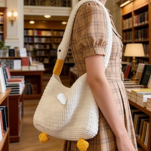 Puede incluir: Bolso de hombro de ganchillo blanco con forma de ganso, con detalles amarillos en el pico y las patas. El bolso tiene una correa blanca y un ala blanca pequeña. La imagen está ambientada en una biblioteca.