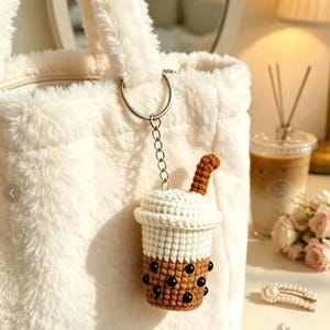 May include: A white faux fur tote bag with a crochet bubble tea keychain attached to the silver ring. The keychain features a brown cup with black beads representing tapioca pearls, a white lid, and a brown straw, all crafted from yarn.