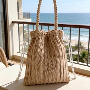 May include: A beige woven drawstring bag with a braided handle. The bag has white drawstrings and is sitting on a white surface. The background shows a balcony overlooking the ocean.