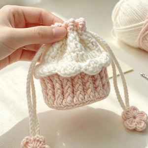 May include: A small, round, crocheted drawstring pouch featuring a light pink base with a vertical ribbed texture and a cream-coloured top section with a scalloped edge. The drawstring cords are cream-coloured and finished with small, light pink flower-shaped accents.