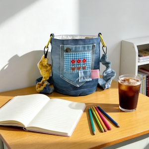 May include: A denim bucket bag with a floral patch and a gingham pocket. The bag has a white interior, yellow and blue straps, and a pink tag. A notebook, pencils, and a glass of iced coffee sit on a wooden desk.