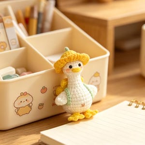 May include: A handmade, white crochet duck wearing a yellow hat and green vest stands next to a beige desk organizer with cartoon graphics. A spiral-bound notebook with lined paper is in the foreground. Pens and stationery are visible in the organizer.