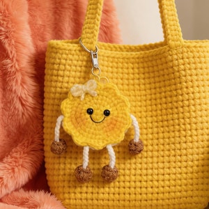 May include: A bright yellow crochet tote bag with matching handles. A smiling, yellow, crocheted sun-shaped keychain with white legs and brown pom-pom feet is attached to the bag. The background includes a peach-colored, fuzzy blanket.
