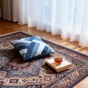 Op de afbeelding: Een kussen met een blauw-wit geometrisch patroon ligt op een patroonkleed. Een kopje thee staat op een klein houten dienblad. Het kleed heeft een gedetailleerd ontwerp met tinten blauw, rood en beige.