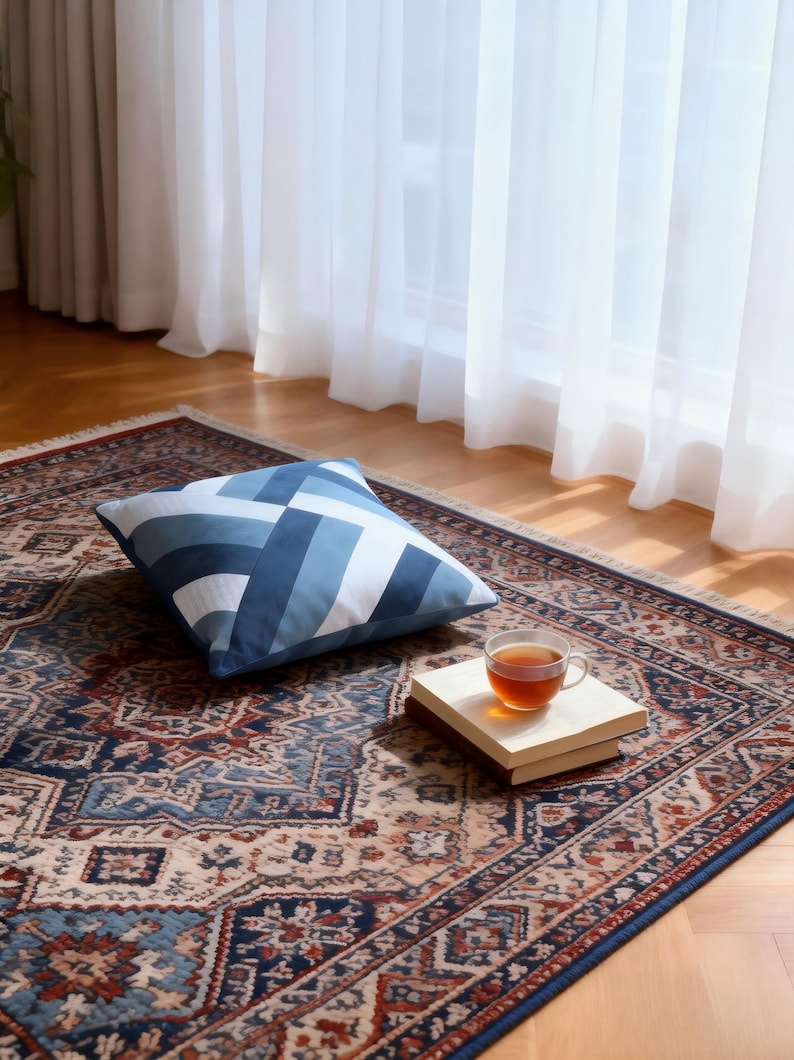 Op de afbeelding: Een kussen met een blauw-wit geometrisch patroon ligt op een patroonkleed. Een kopje thee staat op een klein houten dienblad. Het kleed heeft een gedetailleerd ontwerp met tinten blauw, rood en beige.