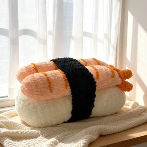 Può includere: Un cuscino di peluche a forma di sushi con una base di riso bianco, un involucro di alghe nori nere e due condimenti di gamberi nigiri. I gamberi sono color pesca con strisce arancioni. Il cuscino è su una coperta di maglia color crema.