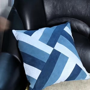 Op de afbeelding: Vierkant sierkussen met een geometrisch ontwerp in blauw- en wittinten. Het kussen heeft een patchworkpatroon met verschillende rechthoekige vormen. Het kussen ligt op een beige oppervlak en een zwarte leren stoel.