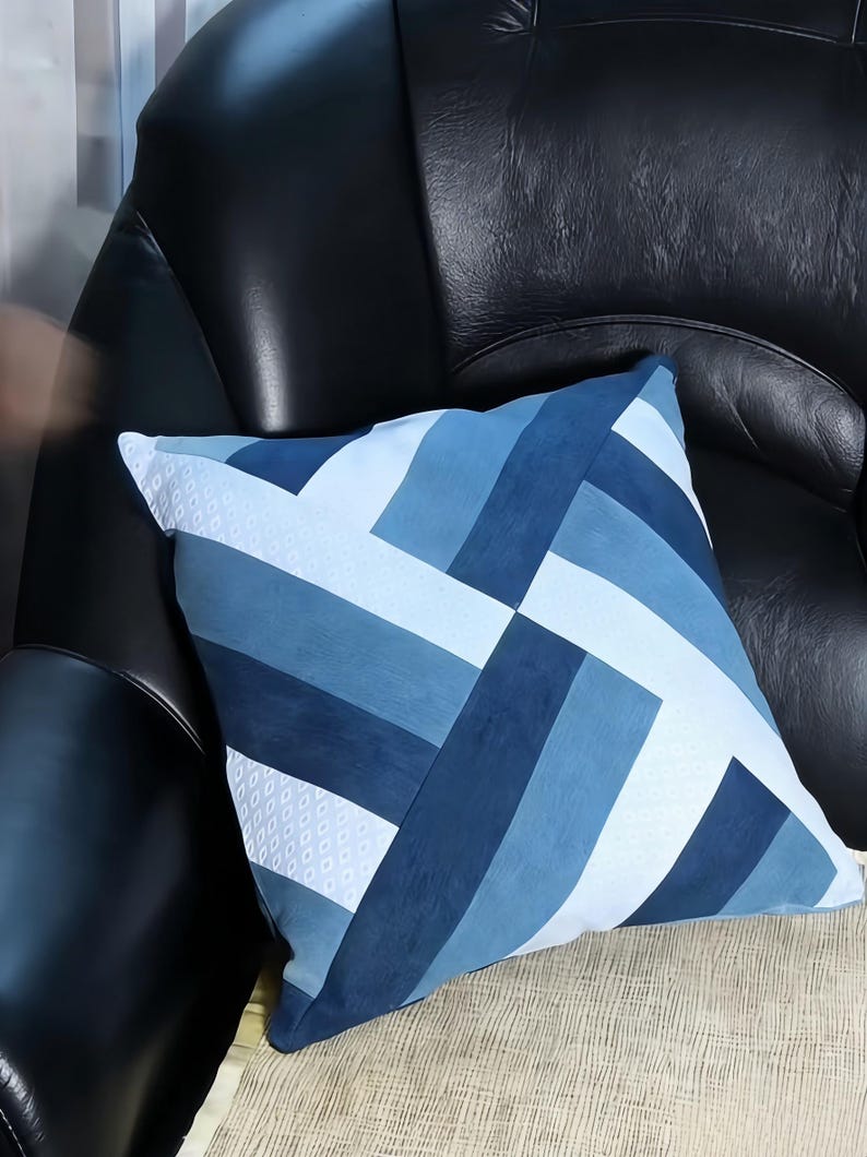 Op de afbeelding: Vierkant sierkussen met een geometrisch ontwerp in blauw- en wittinten. Het kussen heeft een patchworkpatroon met verschillende rechthoekige vormen. Het kussen ligt op een beige oppervlak en een zwarte leren stoel.