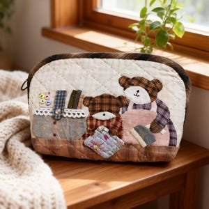 Può includere: Pochette in tessuto trapuntato con un design a orsetto. La pochette presenta uno sfondo trapuntato color crema con accenti a quadri marroni e una chiusura a cerniera. La parte anteriore della pochette presenta orsetti applicati che indossano grembiuli e tengono utensili da cucina.