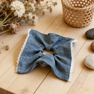 Peut inclure: Un chouchou en jean bleu clair avec une bordure en dentelle blanche. Le chouchou en forme de nœud est posé sur une surface en bois clair, avec un petit panier en osier et des fleurs séchées en arrière-plan.